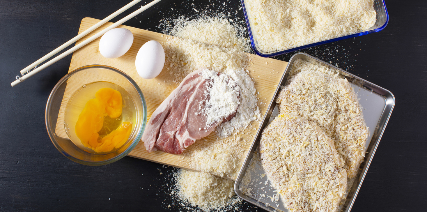 Ingredients for pork cutlet. Eggs, bread crumbs, flour and pork placed on a cutting board.